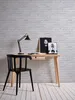 A minimalist desk setup with a black chair, a wooden desk with a lamp and artwork, against a brick wall. Náhled obrázku produktu
