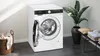 A white Siemens washing machine with its door open, placed in a modern laundry room setting with a basket of clothes and a potted plant. Produktbild-Vorschau