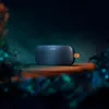 A dark, moody shot of a blue Bose SoundLink Flex speaker with an orange loop, placed on a wooden surface, with blurred foliage in the foreground and background. Produktbild-Vorschau
