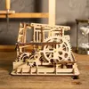 A wooden mechanical model with gears and intricate details, possibly a marble run or automaton, sits on a wooden surface. In the background, a wooden structure and an hourglass are visible. Podgląd zdjęcia produktu