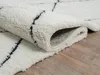 A close-up view of a cream-colored shaggy rug with a black geometric pattern, partially rolled up, resting on a wooden floor. Produktbild-Vorschau