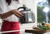 A person in a kitchen holding a pressure cooker, with vegetables on a cutting board in the foreground. Anteprima dell’immagine prodotto