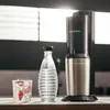 A SodaStream Crystal 3.0 water carbonator is shown on a countertop next to a bottle of water and a glass of sparkling water with strawberries. Produktbild-Vorschau
