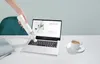 A hand is using a white handheld vacuum cleaner to clean a laptop keyboard. A cup of coffee and some books are on the table. Produktbild-Vorschau