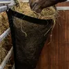 A horse is eating hay from a black mesh hay bag attached to a metal railing in a stable. Anteprima dell’immagine prodotto