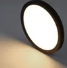 A close-up shot of a black, circular LED ceiling light with a white illuminated center, casting a warm light onto a surface below. Produktbild-Vorschau