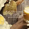 A glass of yellow liqueur is shown on a wooden board, with a basket and a piece of food in the background. Creamy liquid splashes are visible on the left and right sides of the image. Produktbild-Vorschau
