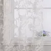A close-up view of a white sheer curtain with an ornate pattern, hanging in front of a window. A small vase with blue flowers is visible on the windowsill. Produktbild-Vorschau
