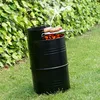 A black barrel BBQ grill is set up on a grassy lawn in front of a dense green hedge, with smoke rising from the grill. Produktbild-Vorschau