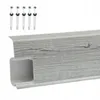 A close-up view of a gray wood-grain patterned PVC skirting board with a white interior channel, along with five screws and wall plugs. Produktbild-Vorschau