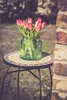 A bouquet of pink tulips in a green glass vase sits on a small mosaic-topped table next to a brick wall. Produktbild-Vorschau