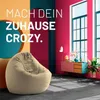 A stylish living room scene with a large beige beanbag chair in the foreground, and a colorful cabinet with decorative items and a plant in the background. Large windows are visible. Produktbild-Vorschau