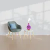 A decorative aroma diffuser emitting mist, placed on a small side table next to a chair, in a well-lit room with shadows on the wall. Voir les images du produit