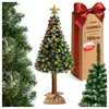 A decorated artificial Christmas tree with a brown trunk and a jute sack base, presented alongside its packaging box and a partial view of another Christmas tree. Produktbild-Vorschau