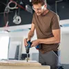 A man is using a cordless drill to drill into a piece of wood. Sawdust is flying from the drilling action. The background shows exposed wiring and ceiling structures, suggesting a construction or renovation environment. Produktbild-Vorschau