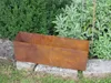 A rusty metal planter box sits on stone blocks in a garden setting, with green bushes and plants in the background. Anteprima dell’immagine prodotto