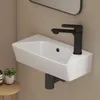 A modern white ceramic wall-mounted sink with a black faucet is shown against a light beige wall. A small potted plant and a bottle are visible on a wooden stool to the left. Anteprima dell’immagine prodotto