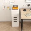 A white filing cabinet with three drawers is shown next to a wooden desk. A desk lamp and an open book are on the desk. A flip calendar and some books are on top of the filing cabinet. Náhled obrázku produktu