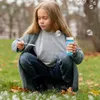 Kind in grauer Strickjacke und Jeans hockt im Gras, hält eine Flasche mit blauer Seifenblasenlösung und einen Blasenstab und bläst Blasen Náhled obrázku produktu
