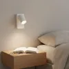A bedside table with an open book and glasses, next to a bed with pillows and a blanket. A wall-mounted lamp is illuminated. Podgląd zdjęcia produktu