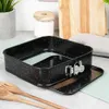 A black springform cake pan with a speckled granite pattern is shown on a countertop with kitchen utensils and a cutting board in the background. Náhľad obrázku produktu