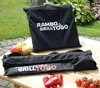 Two black bags with 'RAMBO GRILLTOGO' branding are displayed on a wooden table outdoors, with apples and tomatoes nearby. Produktbild-Vorschau