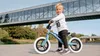 A young boy is riding a blue balance bike outdoors, with a modern bridge structure in the background. Produktbild-Vorschau