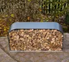 A metal firewood rack filled with logs is placed outdoors on a stone patio, with bushes and a fence in the background. Náhled obrázku produktu