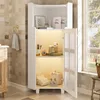 A white corner cabinet with glass doors and shelves filled with bathroom accessories, placed in a bathroom with tiled walls and a window. Produktbild-Vorschau