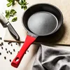A frying pan with a red handle is placed on a light-colored surface, next to a knife, some herbs, and peppercorns. A gray cloth is folded in the foreground. Anteprima dell’immagine prodotto