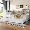 A white daybed with a pull-out trundle bed is set up in a bedroom with a large window and curtains. The daybed has a built-in bookshelf headboard with various items on display. A blanket is draped over the bed. Produktbild-Vorschau