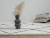 A close-up view of a shaggy rug with a diamond pattern, a vase with dried flowers, and a decorative pillow. Produktbild-Vorschau