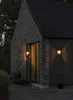 A modern brick and wood house exterior at dusk, with two wall-mounted lights illuminating the entrance. Produktbild-Vorschau