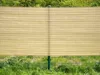 A beige shade screen is attached to a green metal fence, with green grass in the foreground and trees in the background. Produktbild-Vorschau