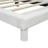 A close-up view of the corner of a white upholstered bed frame, showing the wooden slats of the bed base and one of the cylindrical legs. Produktbild-Vorschau