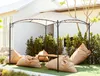 An outdoor seating area with a beige pergola, a wooden table, and several large beige beanbag chairs on a green lawn. Náhled obrázku produktu
