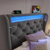 A close-up view of a grey upholstered bed with a blue LED light strip and two electronic devices charging. Náhled obrázku produktu