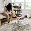 A woman is sitting on a bench next to a baby in a bouncer, reaching out to the baby. Produktbild-Vorschau