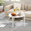 A white round coffee table with storage shelves is placed on a gray rug in front of a beige sofa. The table is decorated with a bowl of fruit, a vase of flowers, and a cup of coffee on a book. Produktbild-Vorschau