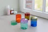 A collection of colorful glass jars with lids, some containing cotton swabs, arranged on a countertop near a window. Produktbild-Vorschau