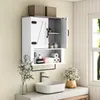 A white wooden bathroom cabinet with two doors, one closed and one open, is mounted on a wall above a sink. The cabinet contains toiletries and toilet paper. A towel bar is attached below the cabinet. A vase with greenery is to the left, and a vase with fl Produktbild-Vorschau
