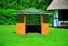 A wooden gazebo with a green roof sits on a green lawn in front of a building with a thatched roof. Produktbild-Vorschau