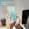 A person is holding a transparent phone case with a smartphone inside, with a laptop and a plant in the background. Produktbild-Vorschau
