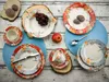 A collection of floral-patterned dinnerware, including plates, cups, and saucers, arranged on a rustic wooden surface with some fruit and stones as decorative elements. Produktbild-Vorschau