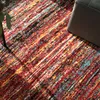 A close-up view of a colorful, abstract patterned rug with a dark gray ottoman in the upper right corner. Produktbild-Vorschau