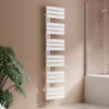 A white towel radiator is mounted on a light brown wall next to a large green plant and a shower enclosure. Podgląd zdjęcia produktu