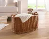 A wicker basket with a blanket and a teddy bear sits on a wooden floor next to a sofa. A window with greenery is visible in the background. Produktbild-Vorschau