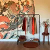 A clothing rack with various garments is placed in front of a large, decorative wall tapestry. A small side table with a vase of flowers and a round rug are also visible. Náhľad obrázku produktu