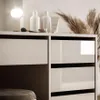 A close-up view of a white dresser with drawers, featuring some cosmetic products and a decorative dried plant on top. Produktbild-Vorschau