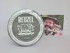 A tin of Reuzel hair pomade is shown next to a postcard featuring a man with a beard and hat. Podgląd zdjęcia produktu
